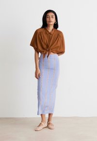 Brune blouse met korte mouwen, geknoopt in de taille, gecombineerd met een lichtblauwe kokerrok met verticale strepen, aangevuld met platte schoenen.
