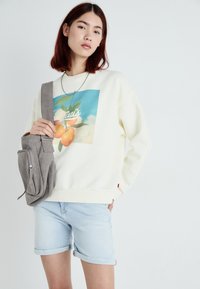 Kremkrāsas džemperis ar krāsainu apelsīnu un lapu grafiku, apvienots ar gaiši ziliem izrullētiem džinsiem un pelēku plecu somu.