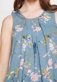 Kukkamotiivinen hihaton paita pehmeässä sinisessä kankaassa, jossa on vaaleanpunaisia ja kultaisia yksityiskohtia, laskoksia keskellä, pyöreä pääntie ja kevyt rakenne.