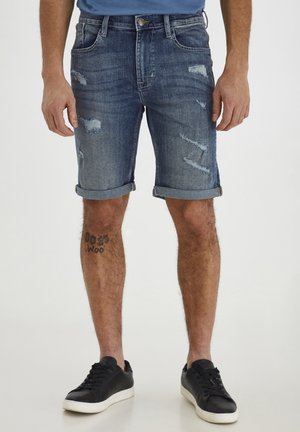 Homme portant un short en jean usé et retroussé ainsi que des baskets noires, debout devant un fond uni.