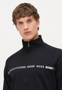 Zwarte zip-up jas met staande kraag, zilveren strepen en het "BOSS" logo op de borst. Zacht materiaal met een gladde textuur.