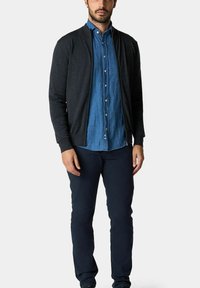 Cardigan grigio scuro con una texture leggera, indossato sopra una camicia di jeans blu con bottoni. Abbinato a pantaloni blu scuro, in piedi contro uno sfondo neutro.
