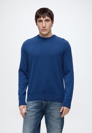 Jeune homme portant un pull bleu uni à manches longues et un jean bleu, debout devant un fond blanc uni.