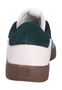 Zapatilla blanca con un talón de ante verde, suela de goma marrón y superficies texturizadas. Presenta un lazo en la parte trasera para facilitar su uso.