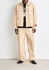 Beige mönstrad jacka med kontrasterande röda sömmar, kombinerad med matchande cargobyxor. Båda har detaljer med fickor och en avslappnad passform.