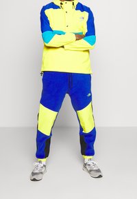 Tuta a blocchi di colore in giallo vivace, blu e nero. Include una giacca leggera a pullover con chiusura a scatto e pantaloni jogger abbinati.