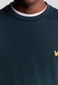 Mörkgrön sweatshirt med ribbad krage. Har en liten gul fågellogo på bröstet. Materialet verkar slätt och texturerat.