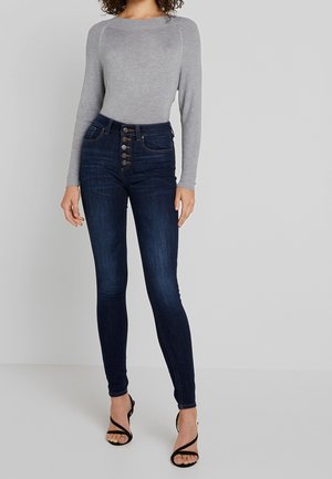 Kvinde iført grå bluse med lange ærmer, mørkeblå højtaljede skinny jeans med knapper foran og sorte stroppesandaler, stående mod en ensfarvet baggrund.