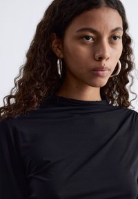 Schwarzes, eng anliegendes Oberteil aus einem glatten, dehnbaren Material, mit einem hohen Halsauschnitt und langen Ärmeln, kombiniert mit großen silbernen Creolen.