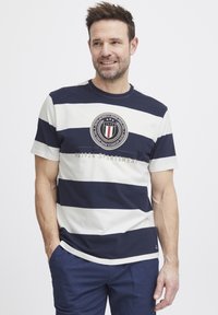Navy and white striped t-paita lyhyillä hihoilla. Rinnassa on pyöreä logo, jossa on tähtiä ja "1924 SPORTSWEAR" -teksti kultaisella.