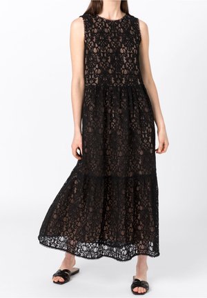 Robe maxi en dentelle noire avec un design sans manches, des motifs floraux et une jupe à volants avec un ourlet en forme de vagues. Portée avec des sandales noires à enfiler.
