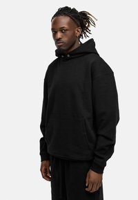 Black hoodie made of soft fabric, featuring a large front pocket, drawstring hood, and ribbed cuffs, worn with matching black pants.
