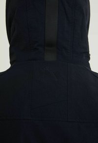 Gros plan sur le dos d'une veste à capuche sombre avec une sangle noire verticale et un logo délicatement brodé sous la capuche.