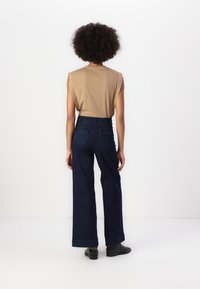 Top bege sem mangas e calças largas de ganga escura de cintura alta, com bolsos traseiros e bainha desfiada. Botas pretas de tornozelo completam o visual.