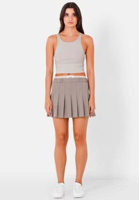 Grauer plissierter Rock mit einem weißen elastischen Bund, kombiniert mit einem beigen, taillierten Tanktop. Das Model steht in weißen Sneakers vor einem weißen Hintergrund.