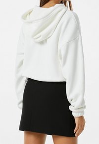 Vit crop hoodie med dragsko huva, mjuk textur och elastiska ärmslut, i kombination med en svart minikjol. Synliga sömnadsdetaljer.