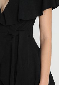 Vestido negro con mangas rizadas sin hombros y un cinturón atado a la cintura, usado por una persona con los brazos al descubierto visible contra un fondo claro.