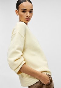 Κρεμ πουλόβερ σε μεγάλο μέγεθος με ριμπ μανσέτες και τελείωμα, από απαλό ύφασμα με υφή. Το μοντέλο στέκεται με τα χέρια στις τσέπες, στραμμένο ελαφρώς πλάγια.