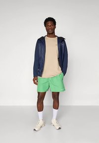 Muž stojaci v námorníckej bunde, béžovom tričku, zelených šortkách, bielych ponožkách a béžových teniskách na jednoduchom svetlom pozadí.