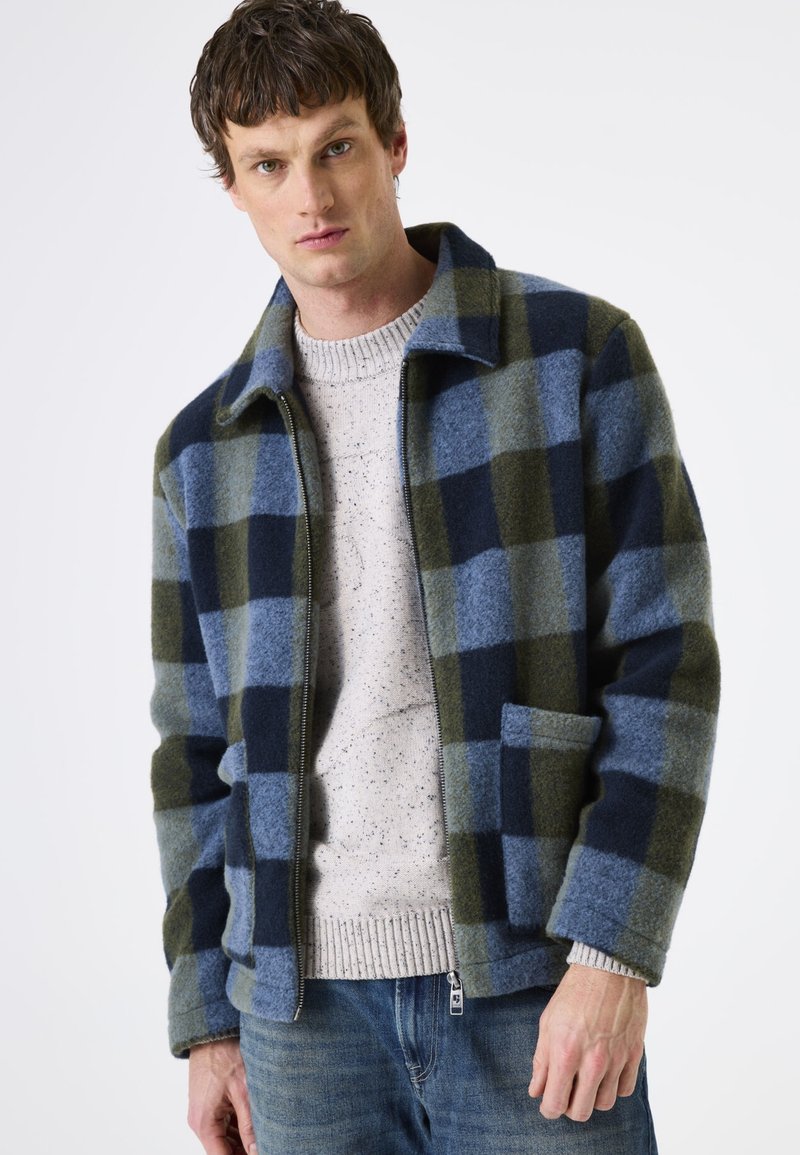 Mann in einer offenen blauen und grünen karierten Jacke über einem hellen gesprenkelten Pullover und blauen Jeans, der vor einem weißen Hintergrund steht.
