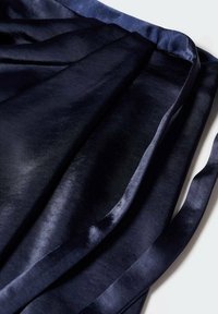 Il tessuto satin navy si drappeggia in morbide e fluenti strati, caratterizzato da una superficie liscia e da una leggera lucentezza. Il bordo ha una rifinitura leggermente più scura per creare contrasto.