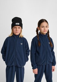 Mörkblå fleecjackor med hög krage, dragkedja framtill och logotillbehör, med matchande byxor, som bärs av två barn.