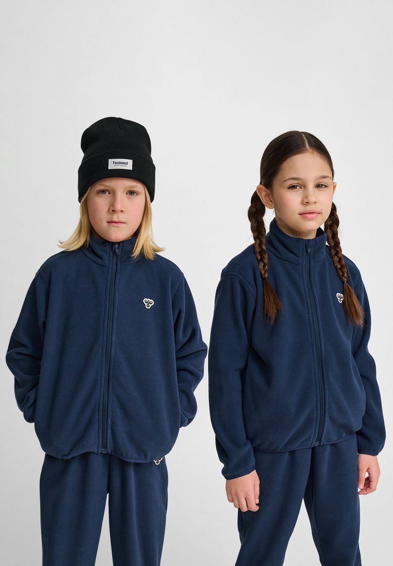 Mörkblå fleecjackor med hög krage, dragkedja framtill och logotillbehör, med matchande byxor, som bärs av två barn.