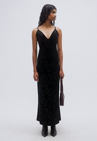 Μακρύ φόρεμα από μαύρο βελούδο με εφαρμοστή σιλουέτα, ντεκολτέ σε σχήμα V, λεπτές τιράντες και λουλουδάτο μοτίβο. Συνδυάζεται με μπότες με τακούνι και σκούρο τσαντάκι.