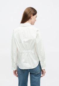 Λευκό πουκάμισ�ο με σφιγμένη μέση, μακριά μανίκια και κουμπιά ως διακοσμητικά στοιχεία. Κατασκευασμένο από απαλό ύφασμα, με κλασικό γιακά και χαλαρή εφαρμογή.