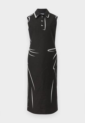 Kobieta w czarnej, bezrękawnikowej sukience midi z białymi abstrakcyjnymi liniami, kołnierzykiem, guzikami i czarnych sandałach na paski, stoi z rękami w kieszeniach.