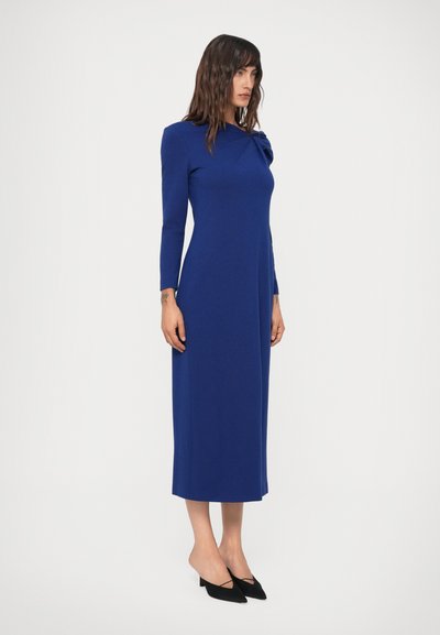 Mulher em um vestido midi de manga longa, azul royal, combinado com sapatos de salto agudo pretos, em um fundo branco liso.