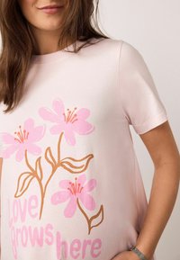 Šviesiai rožinė trumpomis rankovėmis marškinėliai su gėlėtu raštu, kuriame yra rožinės gėlės ir rudi stiebai. Po apačia yra užrašas „Čia auga meilė.“ Minkšta medžiaga.