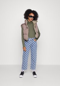 Colete acolchoado em castanho claro sobre uma blusa de gola alta verde, combinado com calças xadrez azuis e brancas e ténis pretos.
