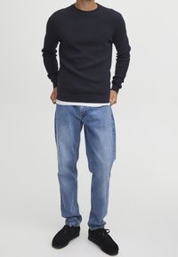 Homme portant un pull en maille bleu marine foncé, un jean bleu clair à jambes droites et des chaussures en daim noir sur un fond blanc uni.