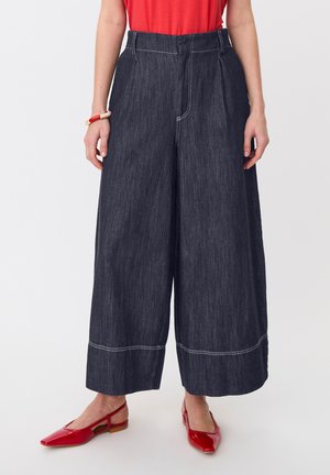 Kvinde iført mørk denim bukser med vidde ben og hvide syninger, rød top, matchende røde slingback sko og et rødt og beige armbånd.