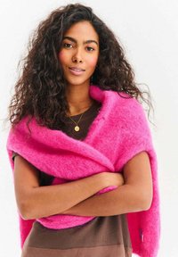 Jasnoróżowy szal wykonany z miękkiego, puszystego materiału, owinięty wokół ramion. Nosi się go na brązowej bluzce, z widoczną metką po prawej stronie.