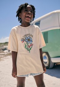 Lys beigefarvet oversized T-shirt med et farverigt blomsterdesign og teksten "VIDUNDERLIG DAG!" sammen med slidte denimshorts.