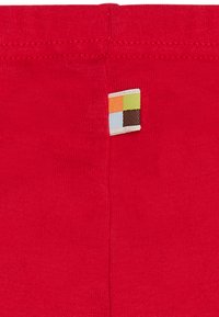Rode stof met een gladde textuur, met een klein, gekleurd geweven label dat aan de bovennaad is gestikt. Eenvoudig ontwerp zonder extra patronen.