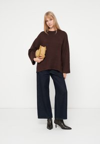 Καφέ πλεκτό πουλόβερ με φαρδιά μανίκια, σε συνδυασμό με σκούρα μπλε φαρδιά παντελόνια και μαύρες μυτερές μπότες μέχρι τον αστράγαλο. Κρατάει μια υφασμάτινη μπεζ τσάντα.