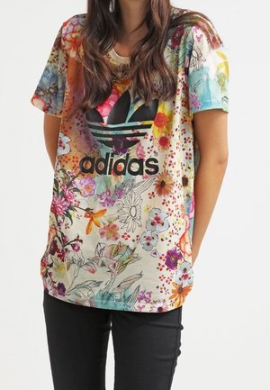 Žena v barevném květovaném tričku Adidas s černým logem, doplněném černými kalhotami, stojí s rukama za zády.