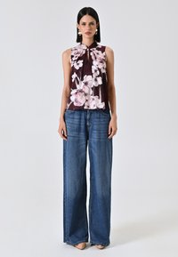 Blusa senza maniche con motivo floreale in un profondo bordeaux; abbinata a jeans a gamba larga blu. La blusa presenta un colletto e un design con pieghe davanti.