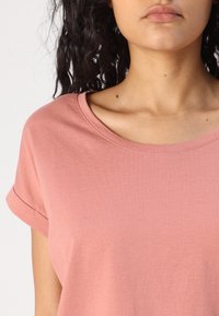 Jasnoróżowa bawełniana koszulka o luźnym kroju, okrągłym dekolcie i podwiniętych krótkich rękawach. Gładka struktura, brak widocznych wzorów lub elementów metalowych.