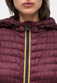 Bordeauxrote Steppjacke mit gesteppter Struktur, einer Kapuze und einem zentralen Reißverschluss mit gelben und blauen Akzenten. Der Stoff wirkt leicht.