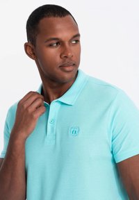 Svetle modré polo tričko z texturované látky, s límečkem, trojicí knoflíků a malým vyšitým logem na hrudi.