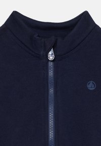 Marinblå zip-up fleecejacka med hög krage, med en kontrasterande dragkedja och en liten logotyp på bröstet. Mjuk textur, avslappnad design.