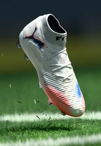Biele kopačky s modrými a červenými akcentmi, v polovici vzduchu nad trávou s bielou čiarou, s čiernym logom Puma na boku.