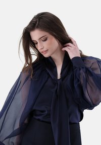 Blusa blu navy con maniche trasparenti e a sbuffo e scollatura annodata. Tessuto liscio con una caduta raffinata e una leggera lucentezza.