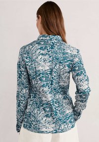 Chemise à manches longues boutonnée en teal avec des motifs floraux blancs, dotée d'un col et d'une coupe décontractée. La vue arrière montre un ourlet incurvé et une texture fine.