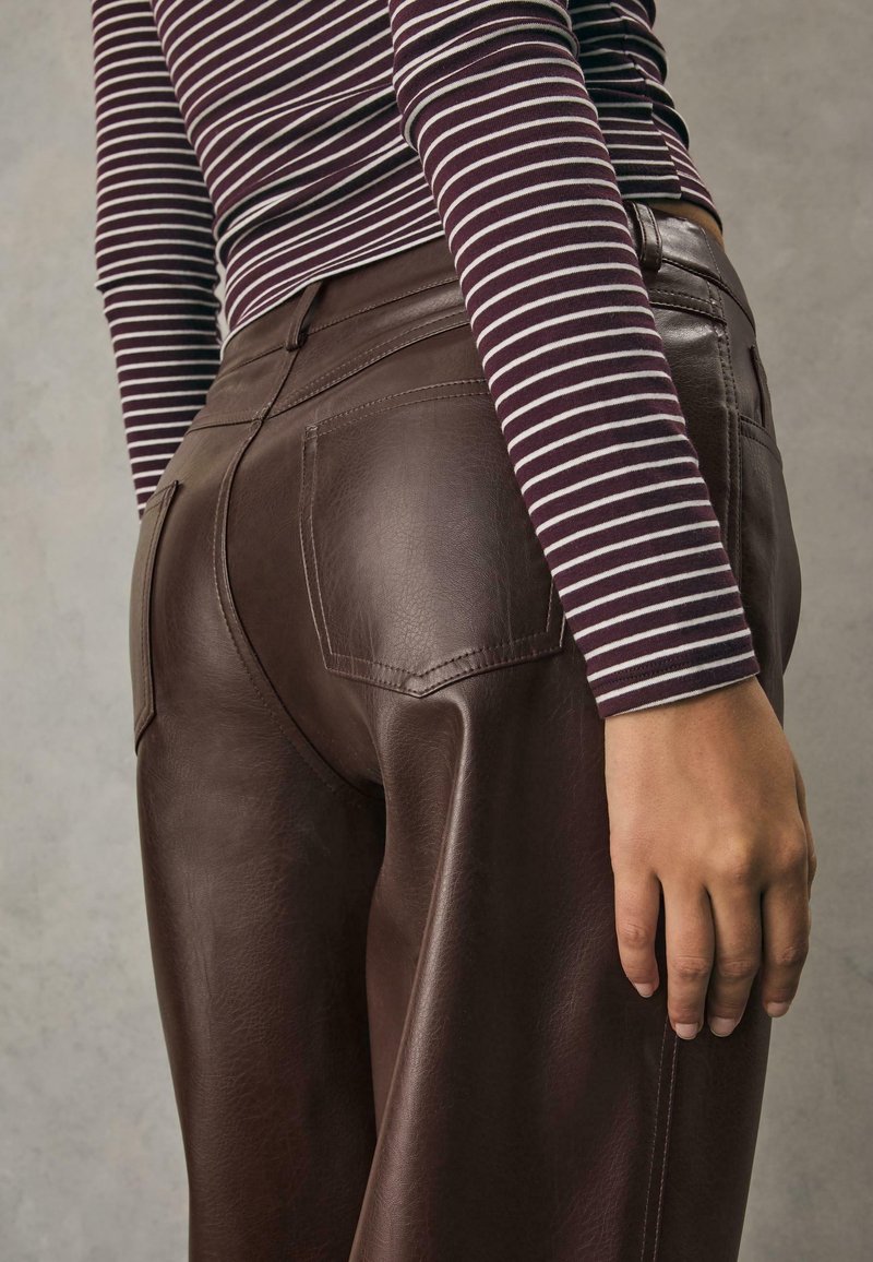 Personne portant un pantalon en cuir marron foncé et une chemise à manches longues à rayures horizontales noires et blanches, debout la main détendue le long du corps.
