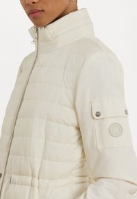 Leichte cremefarbene Puffjacke mit horizontaler Steppung, hohem Kragen und einem Druckknopf-Patch am linken Ärmel mit einem Logo.